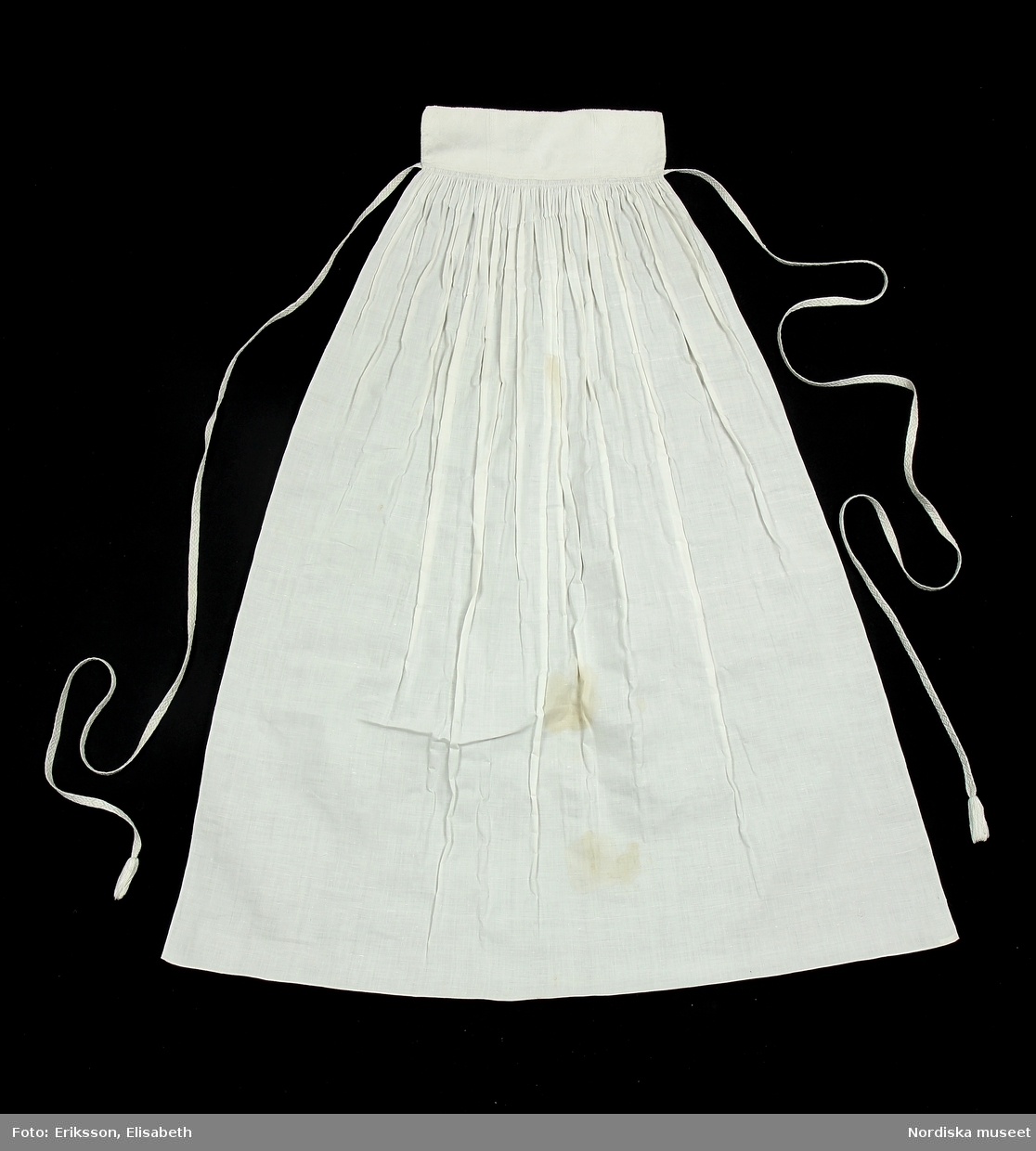 Förkläde för kvinna av tunn, vit, stärkt linnelärft med smal bröstlapp och långa vävda knytband. Tyget är fäst vid bröstlapppen med små, tätt lagda veck, nedtill en smal fåll. Bröstlapp av jacquardvävd damast i halvlinne. Handvävda knytband, 130 respektive 152 cm långa, med mönster i vitt bomullsgarn mot botten av vitt linne, avslutas med ca 4 cm långa tofsar av bomull och lin.

Anm: Flera stora bruntonade fläckar mitt fram.

/2019-09-19 Marianne Larsson
