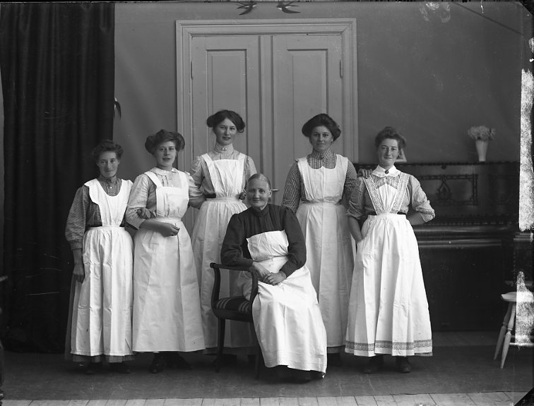 Gruppbild med "jungfrurna på Ribbagården". Fem kvinnor på rad med förkläden står bakom en äldre kvinna med förkläde som sitter på en stol.