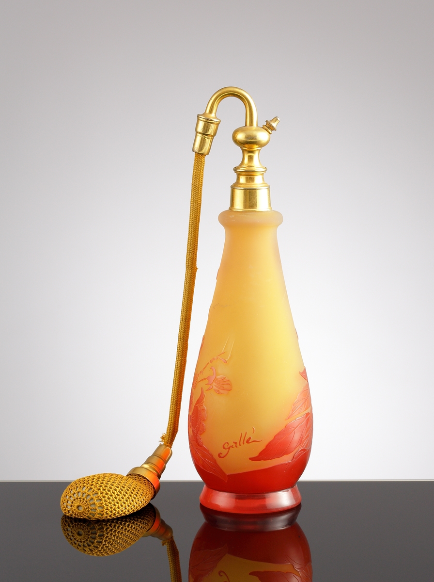Luftfräschare i opakt gult glas med rött etsat överfång. Dekorens motiv är kvistar med blad och blommor. Överst på flaskan sitter en metallfattning med en textilslang försedd med en gummipuff.
