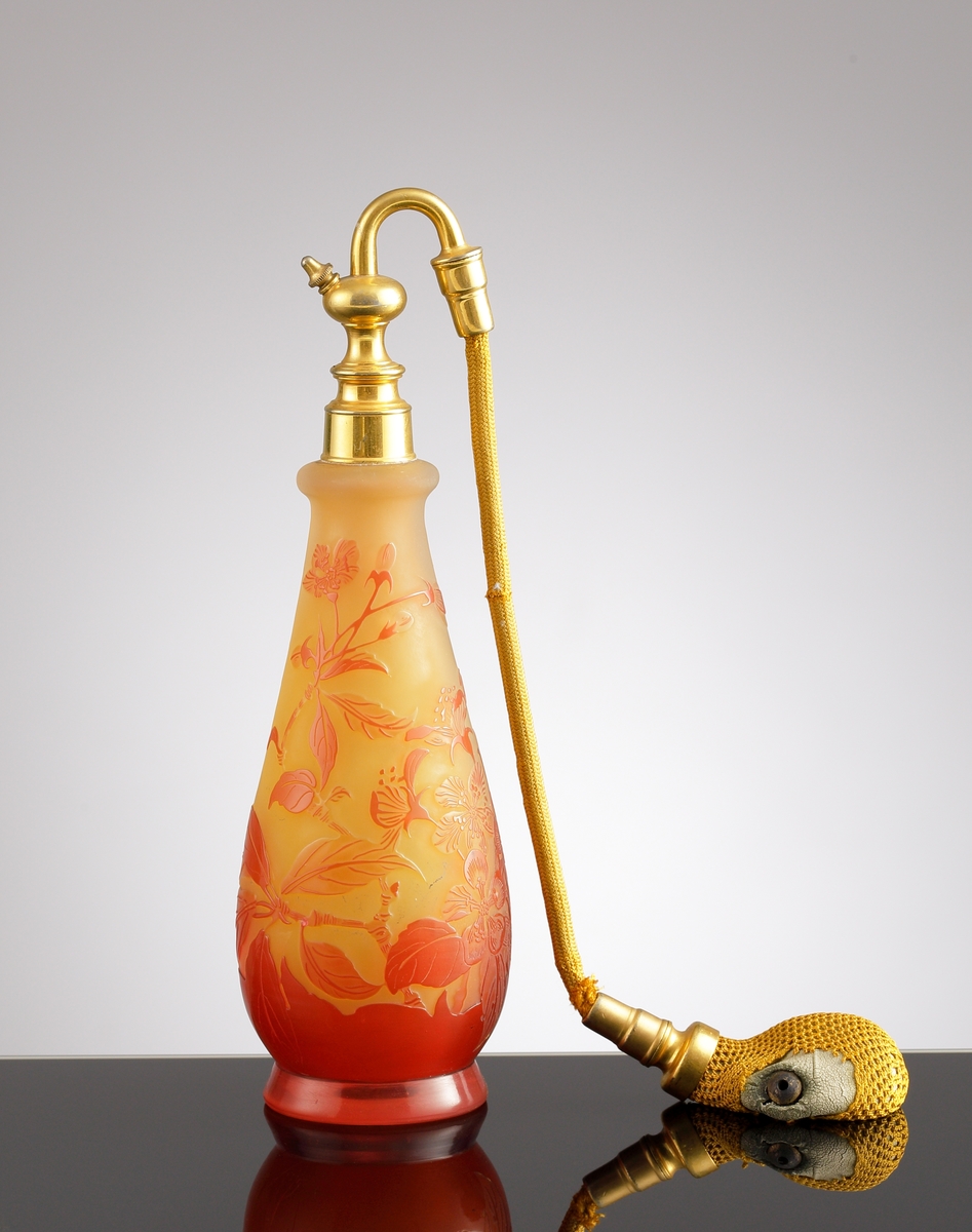 Luftfräschare i opakt gult glas med rött etsat överfång. Dekorens motiv är kvistar med blad och blommor. Överst på flaskan sitter en metallfattning med en textilslang försedd med en gummipuff.
