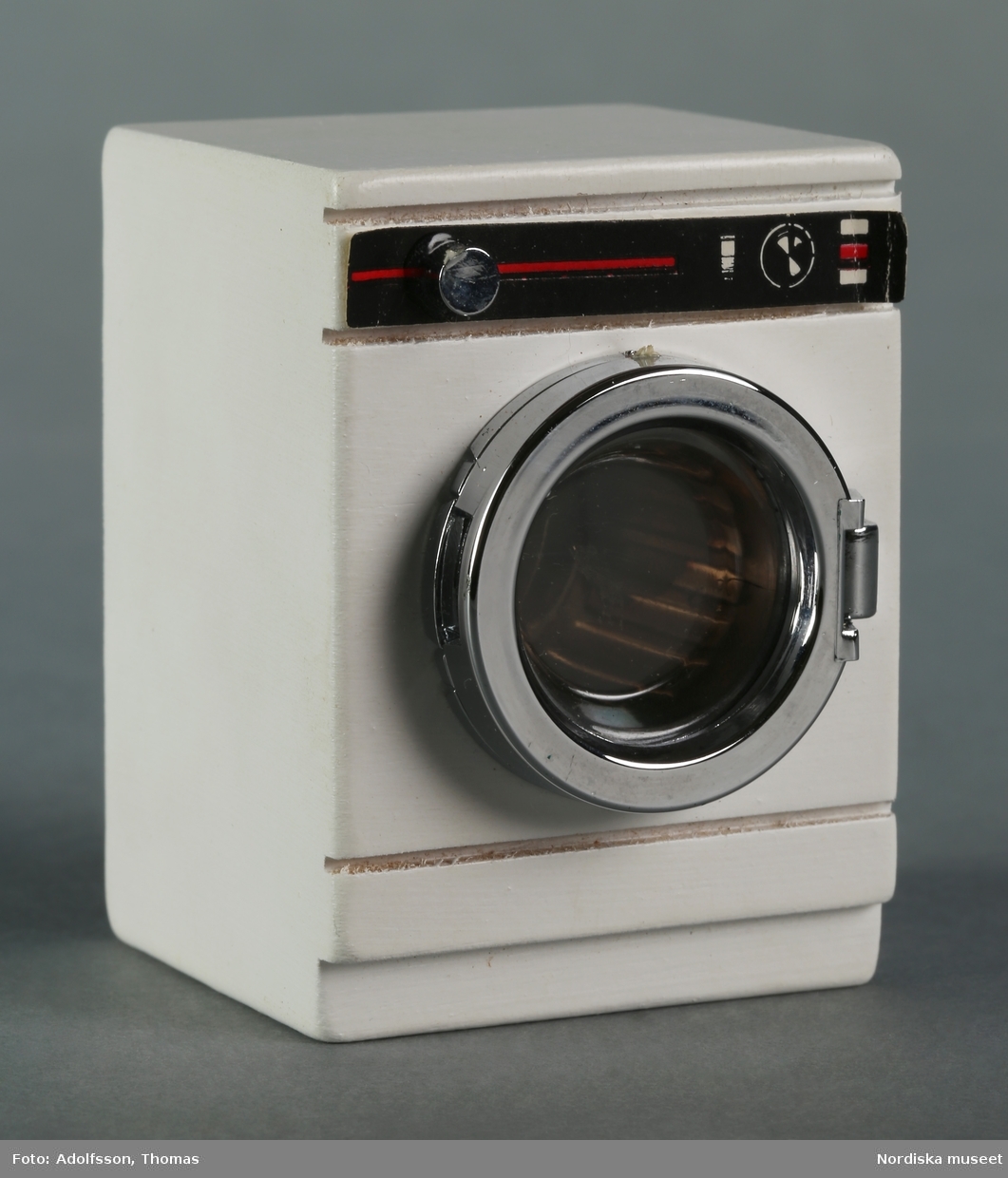 Tvättmaskin av vitmålat trä med tvättrumma av metallfärgad plast samt pappersdekor.som hör till dockskåpsinredningen i källarens tvättstuga i dockskåp NM.0331721+. Till vänster om tvättmaskinen står en tvättlåda (NM.0333456) för förvaring av tvätt och ovanpå maskinen en gul tvättbalja av plast (NM.0333461).