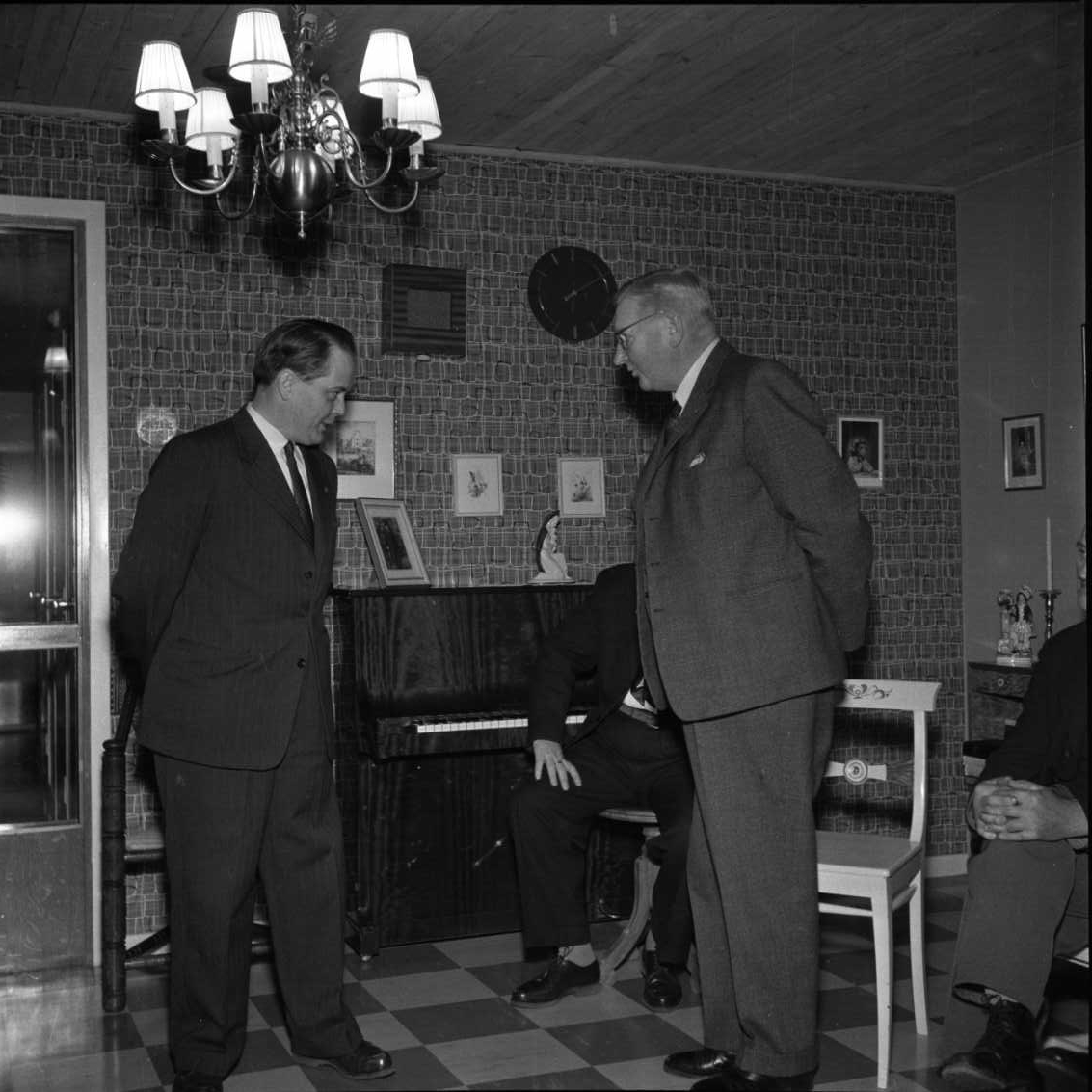 Stadskamrer Bertil Thordow och köpman Gustaf Rahm står mittemot varandra, troligen på Mellbyro ålderdomshem. Den senare avtackas, möjligen avgående i fattigvårdsstyrelsen. I bakgrunden ett piano och skymda personer som sitter på stolar.