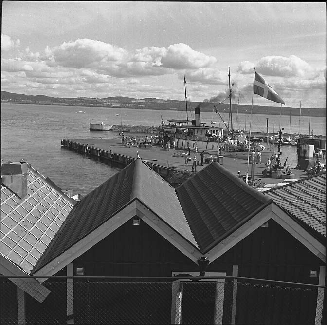 Visingsö hamn, med båthusens tak i förgrunden. Svenska flaggan är hissad vid kaj ligger ångarfartyget Trafik