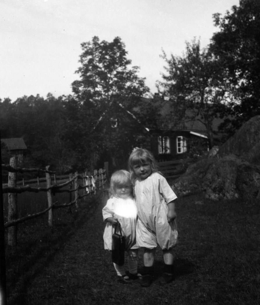 To jenter, trolig to og fire år gammel. En holder ei veske. Bondegård.  Ca. 1920 - 30