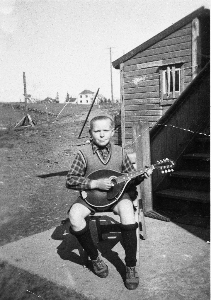 Johannes Stensland (1927 - ) med mandolin. Han sit i bakgarden på faren sin eigendom lengst vest i Storgata. Her er det nå parkering (1990).
Johannes og faren John Juliusson Stensland (1896 - 1972) spela mandolin medan mora Elen Johanne f. Serigstad (1900 - 1976) song.