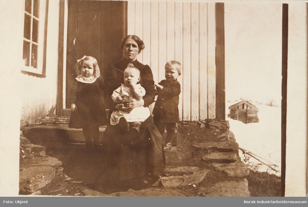 Kvinne med tre barn sittende på trappen foran bolighus
