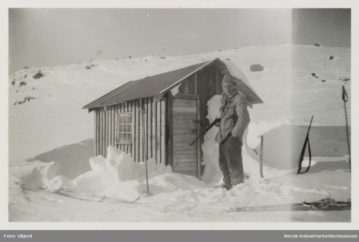 Olav O. Vågen står med gevær i snøen foran jakthytta Olavsbu i Kvamsfjellet