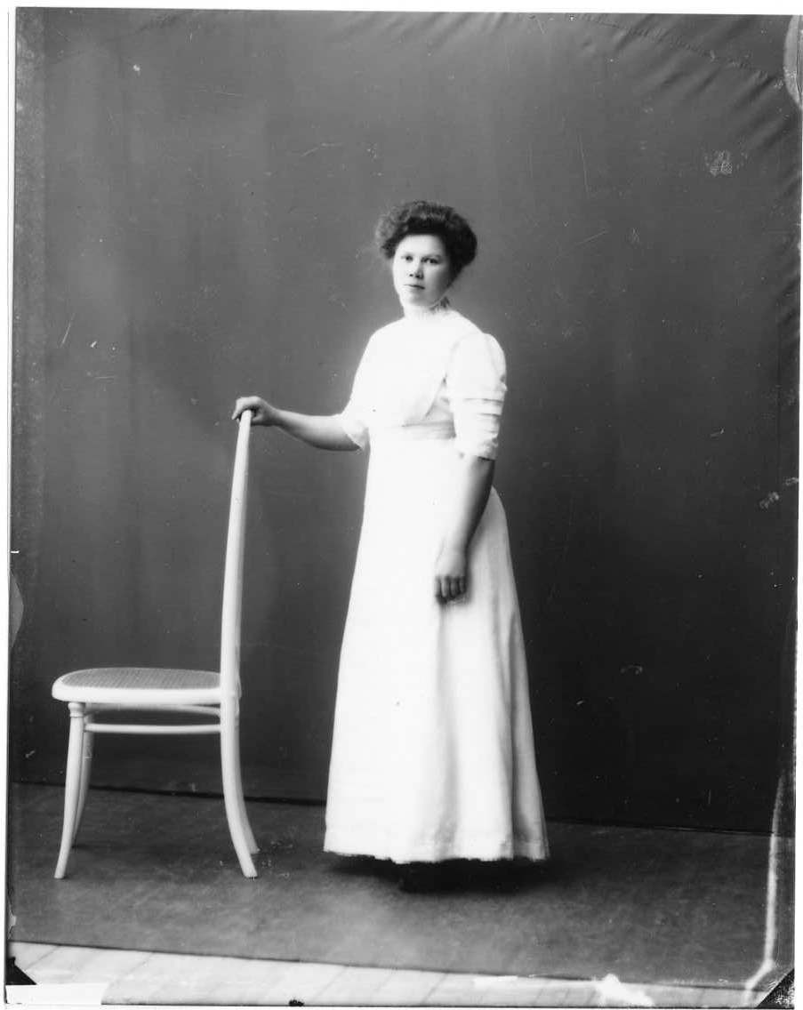 Porträtt av Rut Andersson från Örserum. Hon står i en fotsid ljus klänning med höger hand vilande på en stolsrygg.