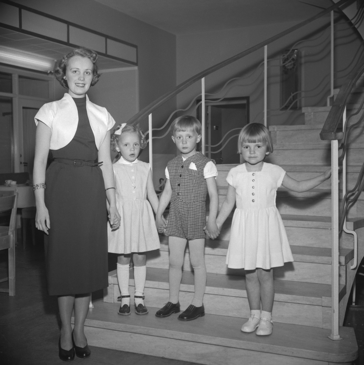Motiv från modevisning i Ljungsbro 1954. Klänning med skärp och pennkjol till klackskor låg helt i tiden, här kompletterad med en kortärmad bolero. Barnen bär exempel på lediga plagg som vid tiden slår igenom. Platsen är ortens kommunhus.