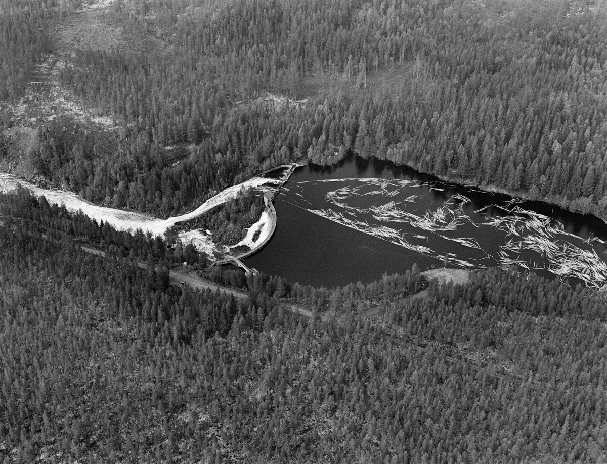 Flyfotografi tatt over kraftverksdammen i Osfallet i elva Søndre Osa i Åmot i Hedmark i mai 1984, den siste sesongen det foregikk tømmerfløting i denne delen av Glomma-vassdraget.  Fotografiet viser hvordan tømmerstokkene kom flytende med strømretningen over kraftverksdammen, der det var lagt ut lenser for å lede virket mot tømmerløpet som var plassert mot det nordre landet, hvor fallhøyden var mindre enn nedenfor den buete hoveddammen mot søndre landside.  Etter å ha passert dammen ble stokkene ført med strømmen i kvitskummende vann ned mot utløpet i elva Søndre Rena, cirka to kilometer lengre vest. Damanlegget på fotografiet skal ha vært ferdigstilt i 1947.  Den ble noe ombygd i 1981.  Ettersom vassdragslovgivinga påla dem som drev tømmerfløting å fjerne installasjoner i vann ved opphør av virksomheten, ble tømmerløpet ved Osfallsdammen revet umiddelbart etter at fløtinga på dette fotografiet var avviklet.  Energiproduksjonen ved denne lokaliteten foregår i en kraftstasjonsbygning fra 1914, cirka en kilometer nedenfor den avbildete dammen, hvor det er installert to francisturbiner som kan yte 5.5 megawatt.  Den opprinnelige dammen ble ødelagt ved et dambrudd i 1916.  Det nåværende kraftverket utnytter et fall på 41 meter i elva Søndre Osa.  Vegen på sørsida av vassdraget fører ned til kraftstasjonen og derfra mot Fylkesveg 215 ved Oset.