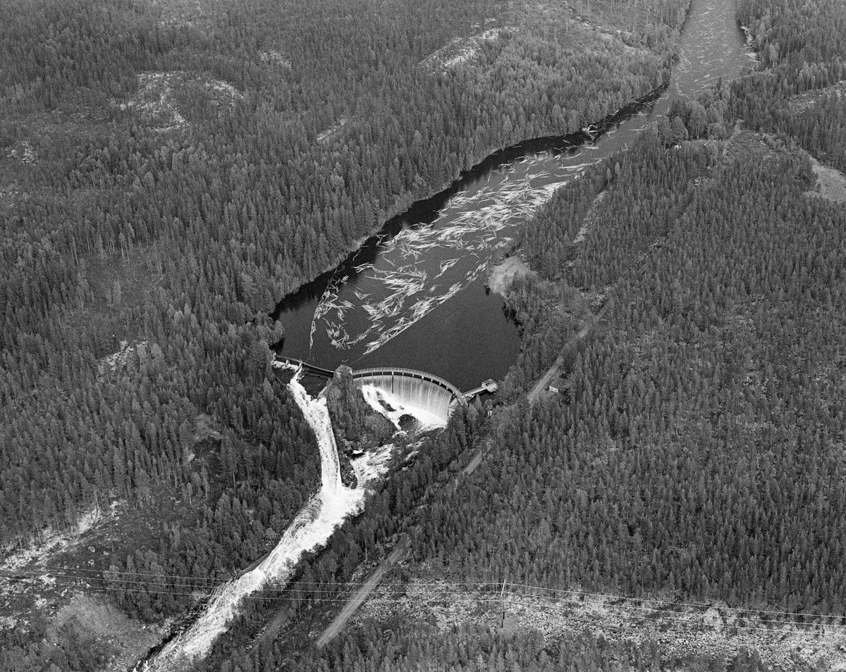 Flyfotografi tatt over kraftverksdammen i Osfallet i elva Søndre Osa i Åmot i Hedmark i mai 1984, den siste sesongen det foregikk tømmerfløting i denne delen av Glomma-vassdraget.  Bildet er tatt fra sør.  Fotografiet viser hvordan tømmerstokkene kom flytende med strømretningen over kraftverksdammen, der det var lagt ut lenser for å lede virket mot tømmerløpet som var plassert mot det nordre landet, hvor fallhøyden var mindre enn nedenfor den buete hoveddammen mot søndre landside.  Etter å ha passert dammen ble stokkene ført med strømmen i kvitskummende vann ned mot utløpet i elva Søndre Rena, cirka to kilometer lengre vest.  Damanlegget på fotografiet skal ha vært ferdigstilt i 1947.  Den ble noe ombygd i 1981.  Ettersom vassdragslovgivinga påla dem som drev tømmerfløting å fjerne installasjoner i vann ved opphør av virksomheten, ble tømmerløpet ved Osfallsdammen revet umiddelbart etter at fløtinga på dette fotografiet var avviklet.  Energiproduksjonen ved denne lokaliteten foregår i en kraftstasjonsbygning fra 1914, cirka en kilometer nedenfor den avbildete dammen, hvor det er installert to francisturbiner som kan yte 5.5 megawatt.  Den opprinnelige dammen ble ødelagt ved et dambrudd i 1916.  Det nåværende kraftverket utnytter et fall på 41 meter i elva Søndre Osa.  Vegen på sørsida av vassdraget fører ned til kraftstasjonen og derfra mot Fylkesveg 215 ved Oset.