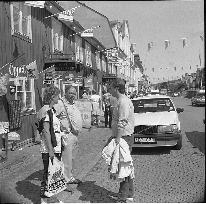 Sigge Johansson står med okänd kvinna och man på trottoaren utmed Brahegatan i Gränna. Bakom dem står en Volvo parkerad. På husfasaden hänger ett stor antalt vimplar med "Öppet", "Vivo" etc