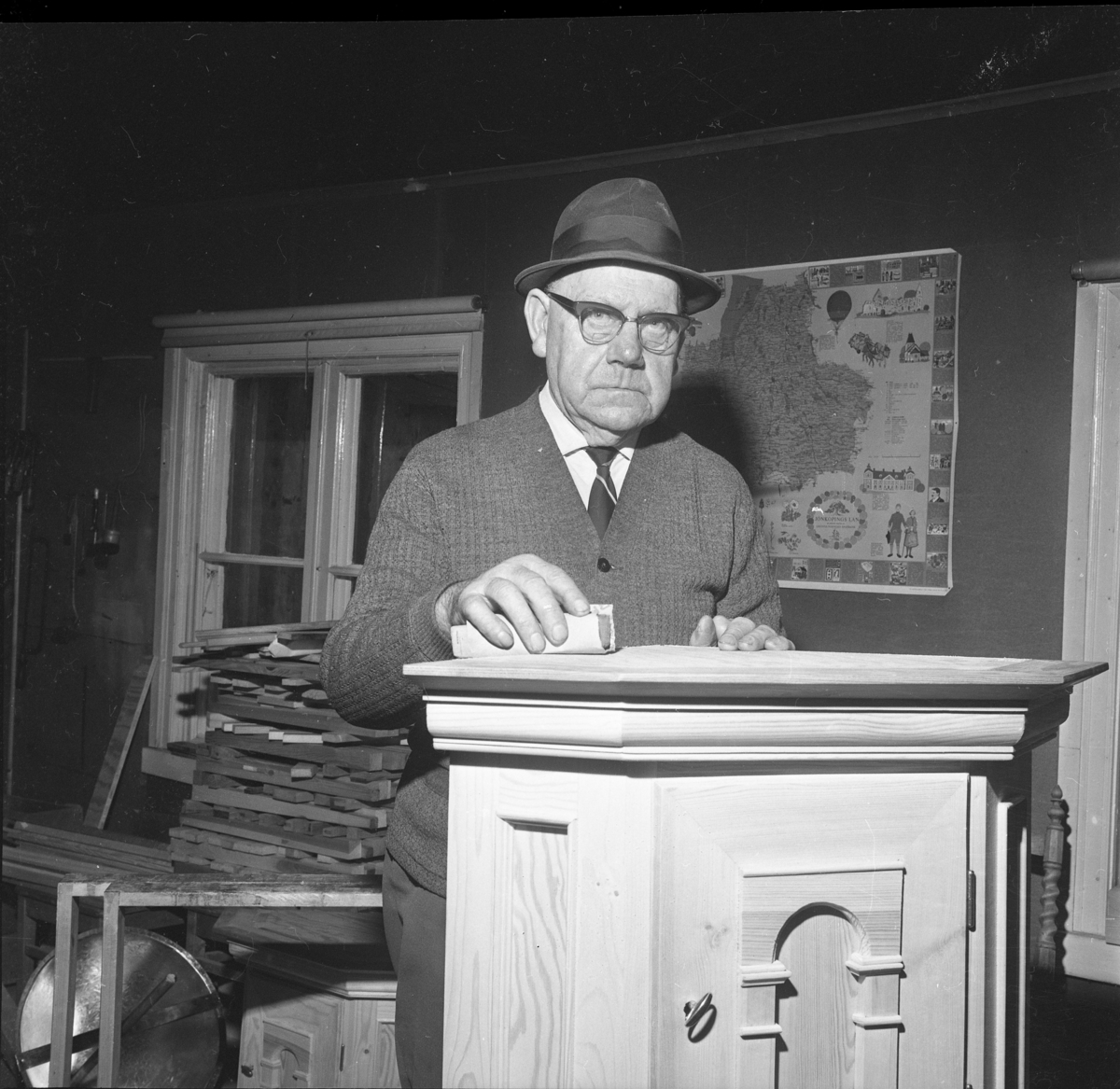 Vaktmästaren och snickaren Gunnar Berglund, med glasögon och hatt på huvudet, står troligen i en snickarbod med ett skåp som han har tillverkat.