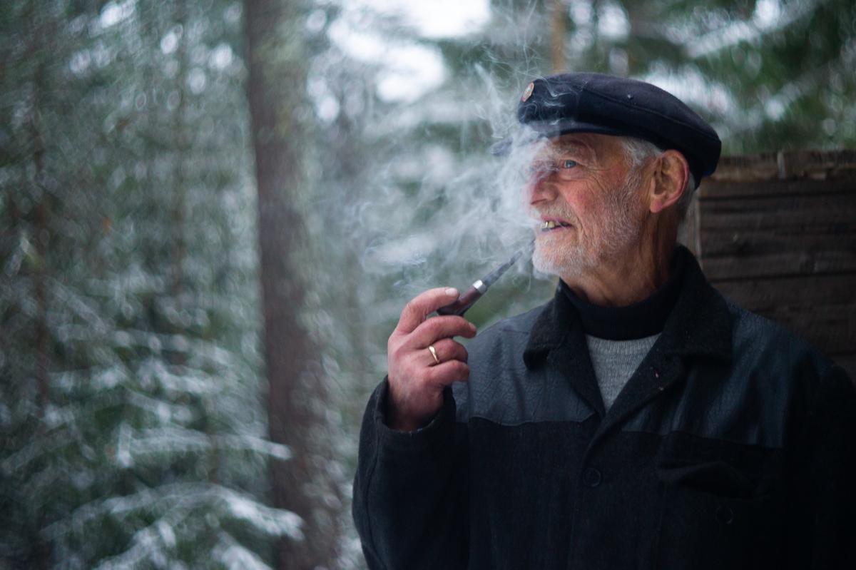 Fotografiet viser en kar med pipe. Han står utenfor tømmerkoia på Odalstuner. Foto er tatt under Jul i stuene, og det er snø på trærne i bakgrunn.