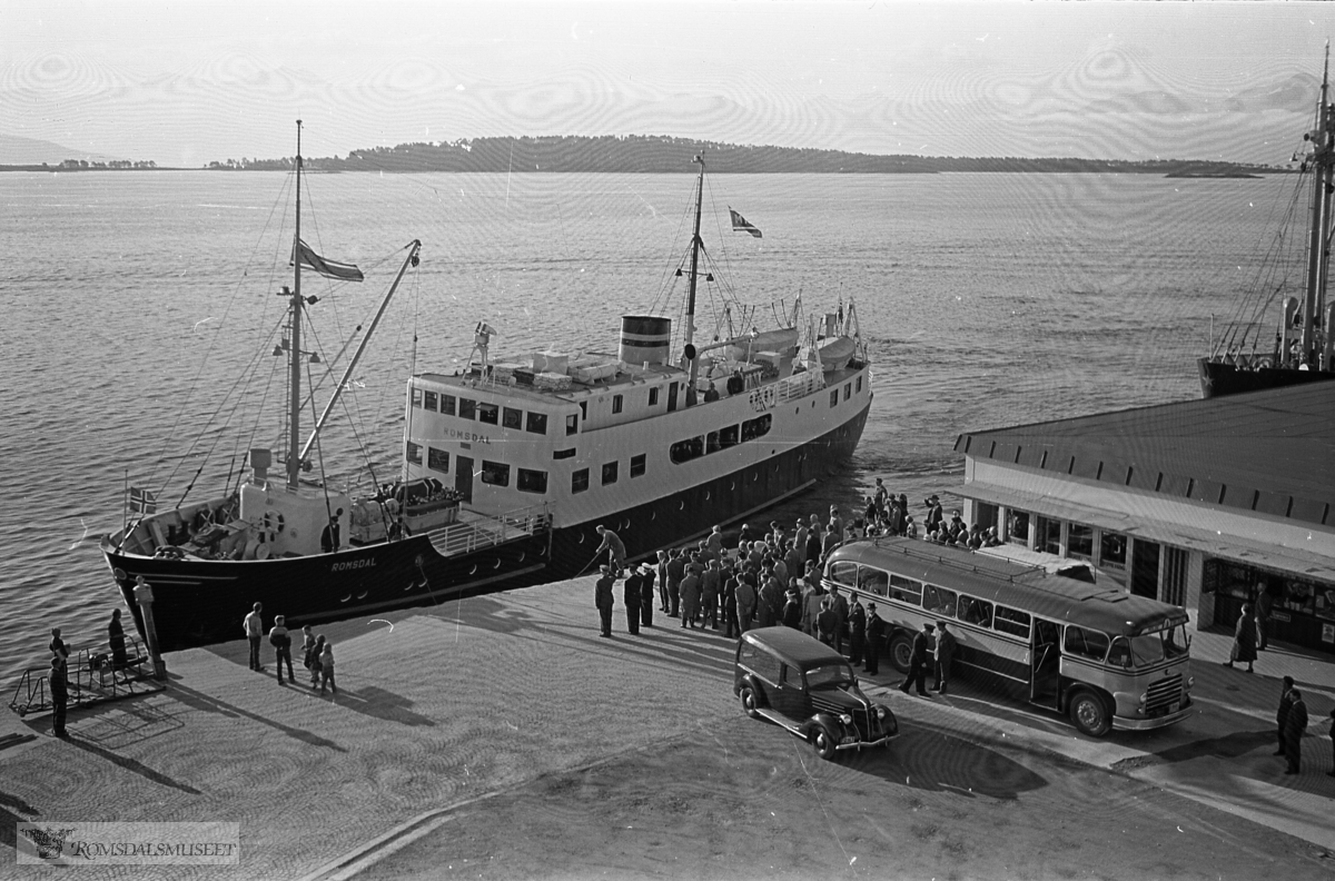 "September 1958"."Oksviks gravferd".Fra begravelsen til Olav Oksvik f.07.05.1887 d.16.09.1958..Båten som legger til kai er M/S Romsdal, som gikk i rute Åndalsnes-Molde-Ålesund i tidsrommet 1950-1962. Den ble solgt i 1962..Bussen på bildet er sannsynligvis T-8685 Scania-Vabis, 1953-modell, med karosseri fra Bussbygg, Hovdenakken. Bussen tilhørte Aarø Automobilselskap (Auto) og den var lakkert i gult og mørk blå. Aarø Auto hadde ruter Molde-Kristiansund over Gjemnes, og ruta startet og avsluttet ved Storkaia i Molde, der det var korrespondanse med båter til Åndalsnes og Ålesund. .Begravelsesbilen er en Ford V8 1936 med registreringsnummer T-2167.