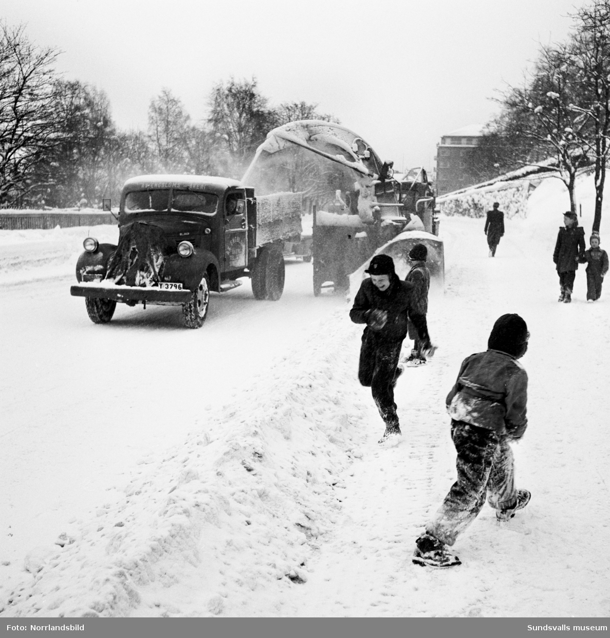 Snöröjning med plog, lastbilar och snöslunga på västra Storgatan. Några barn leker en farlig lek framför snöslungan.