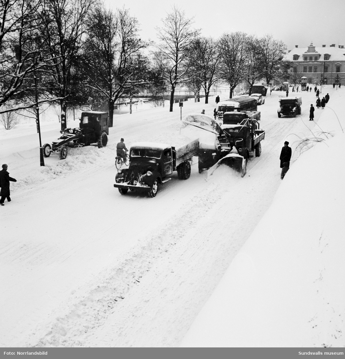 Snöröjning med plog, lastbilar och snöslunga på västra Storgatan. Några barn leker en farlig lek framför snöslungan.