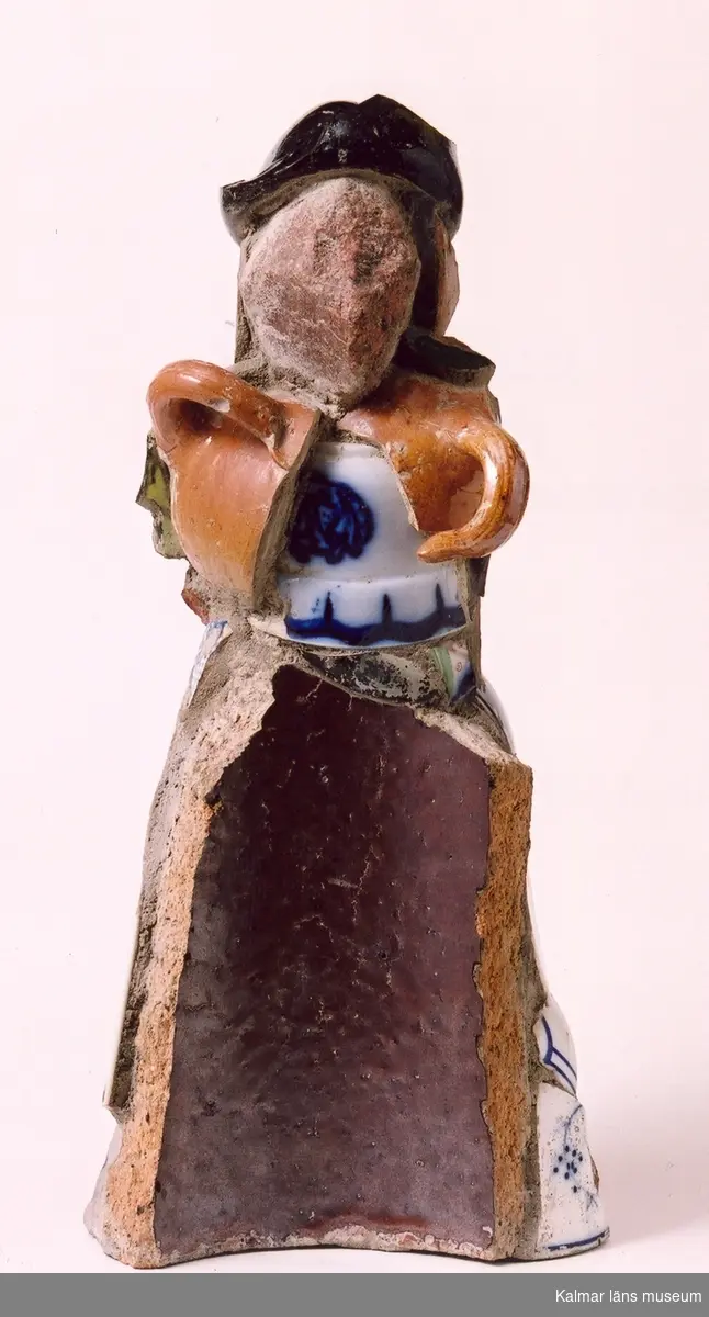 KLM 28161:1 Skulptur, collage med skärvor av diverse keramik på stomme av betong. Modellnamn: Måna-Lisa. Signerad: O. Nyman -63. (Olle Nyman, 1963). Kvinnofigur sammansatt av lergods, porslin, glasbitar samt en sten som ansikte. Så utgörs t ex kjolens framsida av ett stycke saltglaserat dräneringsrör, baksidan delar av stora blomkrukor, armarna är hänklar till geneverkrus och hatten av botten till en grön butelj med Kinnekulle, dessutom bitar av mönstrat porslin. 
Av givaren avsedd som länsmuseets maskot (beskyddare av keramiksamlingarna) och döpt till "Måna-Lisa" - vi ska "måna om" den.