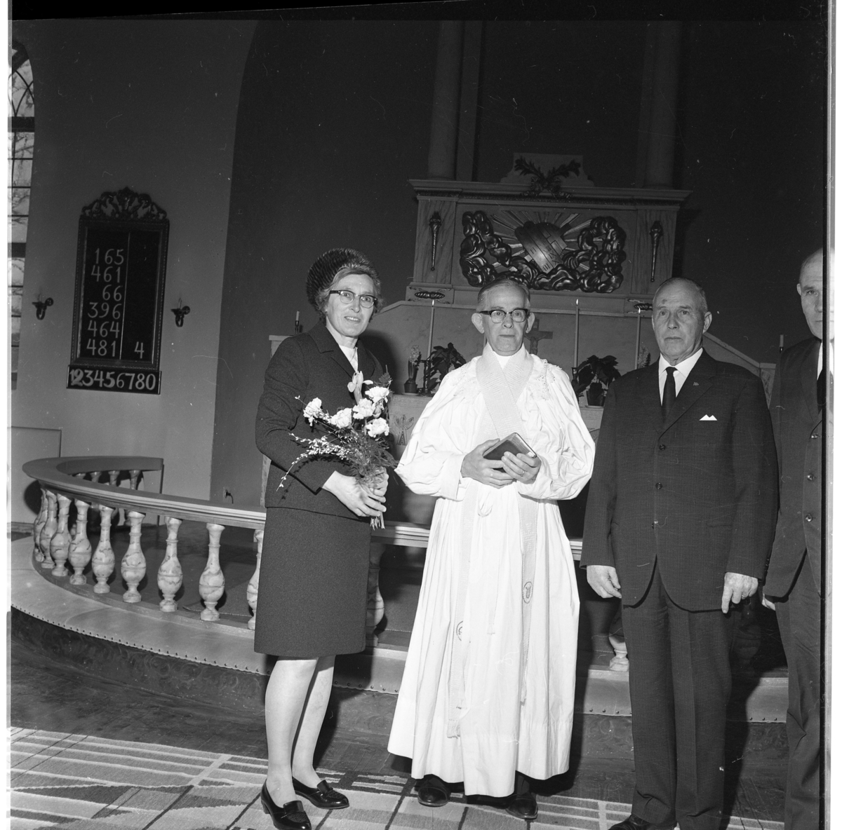Fröken Helena Johansson med blommor i handen står intill kyrkoherde Josef Lönnesten och kyrkvärdarna Hugo Carlsson och Karl Fritzell. De står framför altaret i Adelövs kyrka.