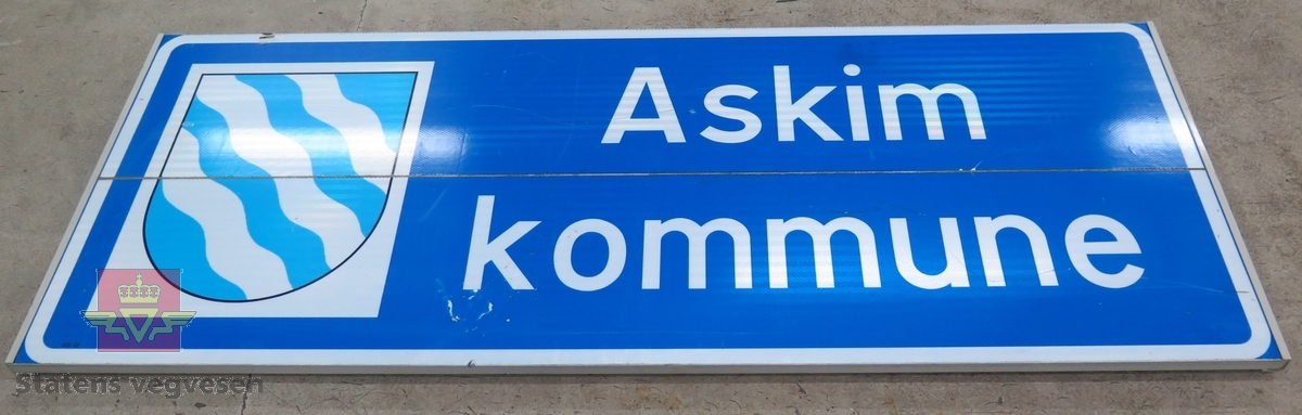 Flerfarget skilt av aluminium der to plater er skrudd sammen med klammer. Skiltet er lysreflekterende, foliert. med kommunevåpen og har teksten: Askim kommune.
