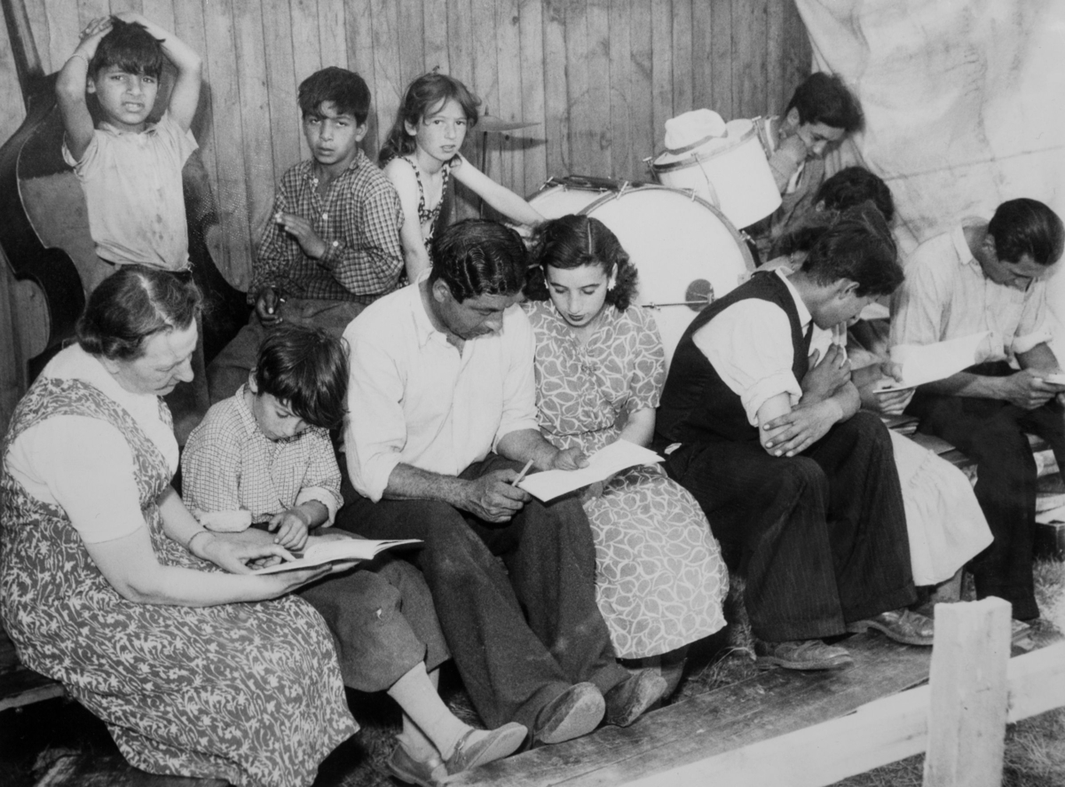 En grupp romer i olika åldrar sitter och läser under sommarskola 1950. Längst till vänster sitter fröken och hjälper en yngre pojke.