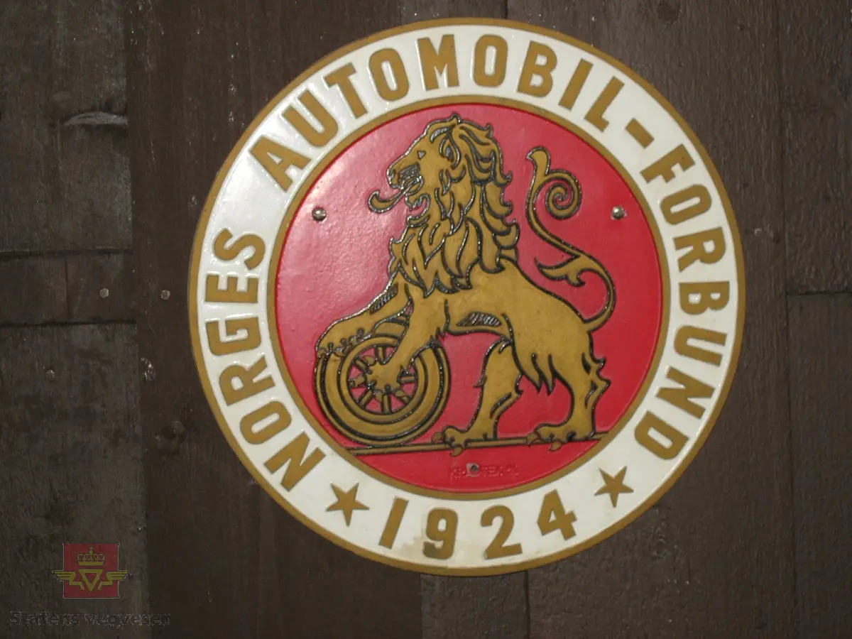 Rektangulært navneskilt i metall, med teksten  "NORGES AUTOMOBIL-FORBUND 1924" med gullfargede bokstaver på rød/hvit bakgrunn, med NAF's logo i midten.