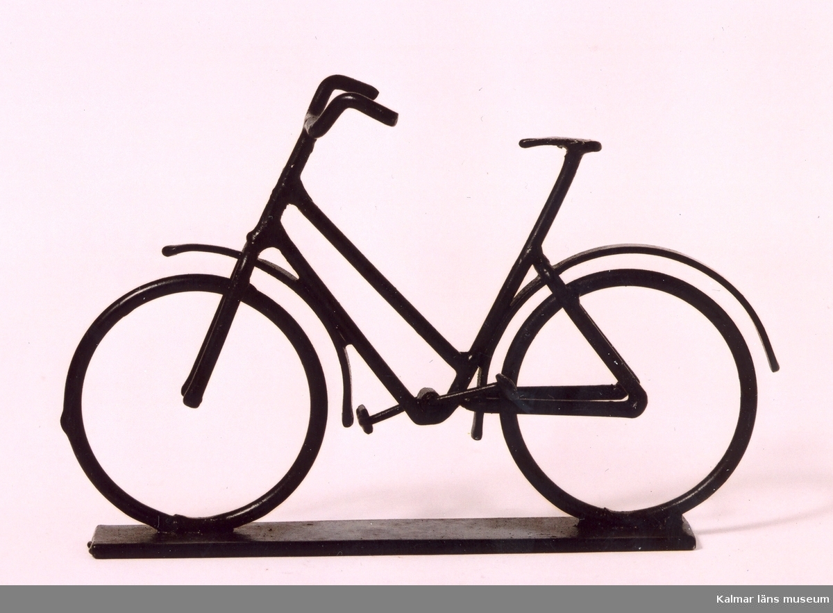 KLM 39586:48:1-2 Cykel med upphängningsanordning i form av en ram för att hängas på väggen, av metalltråd, svartmålad. Cykeln, monterad på metallplatta. Tillverkad i konstsmide, svetsad. Damcykel med vanligt styre. Cykeln är inte monterad i den medföljande ramen. Prydnadsföremål. Tillverkad av Yngve Axtelius som var verksam som trådslöjdare.