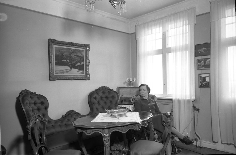 Fröken Astrid Malmborg sitter i en fåtölj vid en soffgrupp intill fönster med ljusa, tunna gardiner. Det är hennes föräldrars hem. På väggen mellan fönsterna några ramade fotografier, bland annat en danspose med henne själv. På väggen bakom soffan hänger en målning av hennes pappa, konstnären Pelle Malmborg, med ett motiv från Gränna.