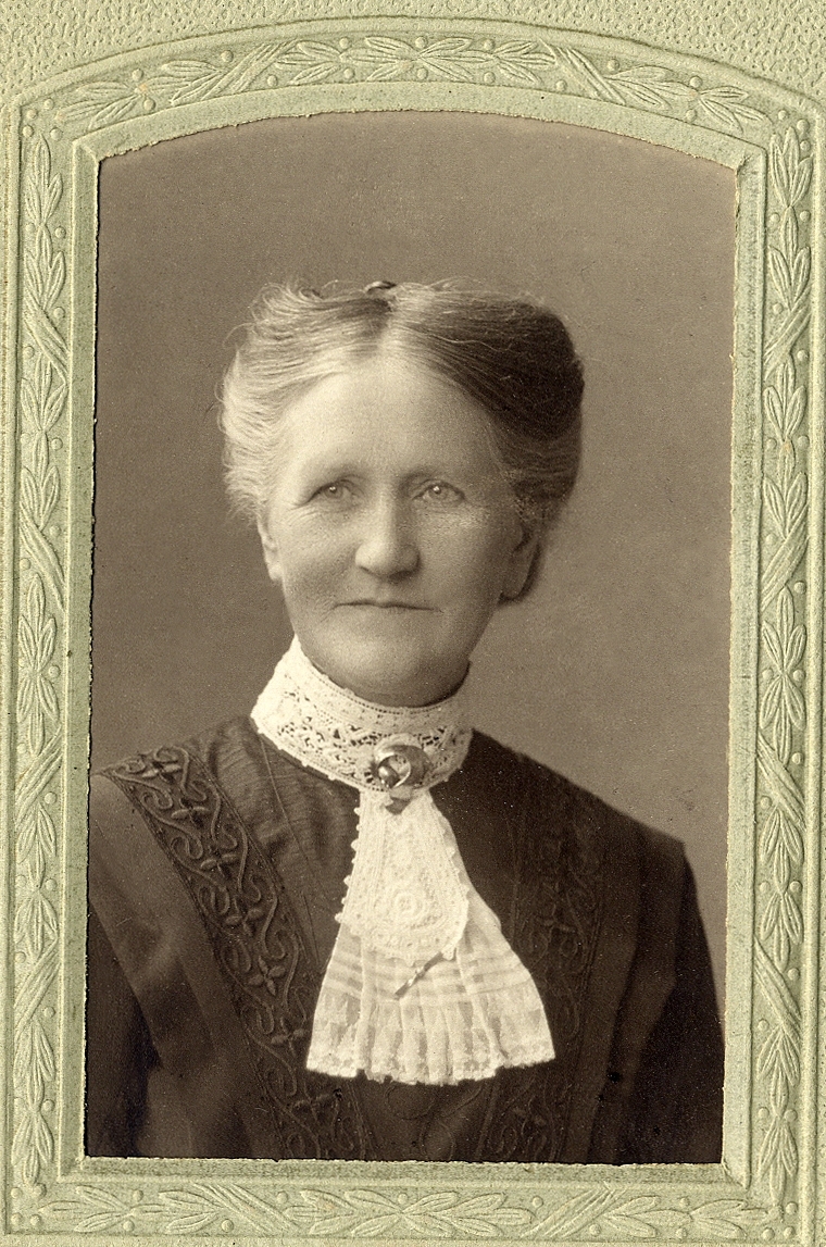 En okänd äldre kvinna i mörk klänning med hög vit spetskrage och spetsfischy. 
Vid kragen syns en brosch och längre ner en slags "slipshållare" i form av en smal brosch. 
Bröstbild, halvprofil. Ateljéfoto.