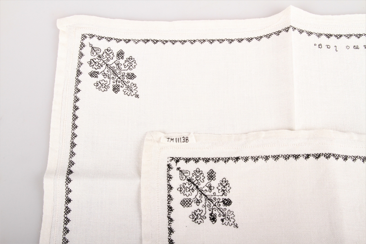 Bordduk med hvit bunn og med svart og hvit dekor. Brodert med korssting, kontursting og hullsøm, border og blomster samt tekst.