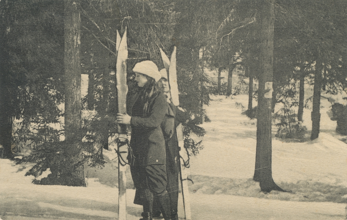 Postkortmotiv av to kvinner som holder sine ski i snødekt landskap.