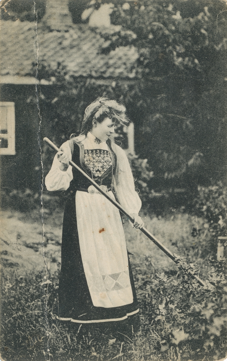 Postkortmotiv av ei kvinne i bunad som står i gresset og holder et redskap.