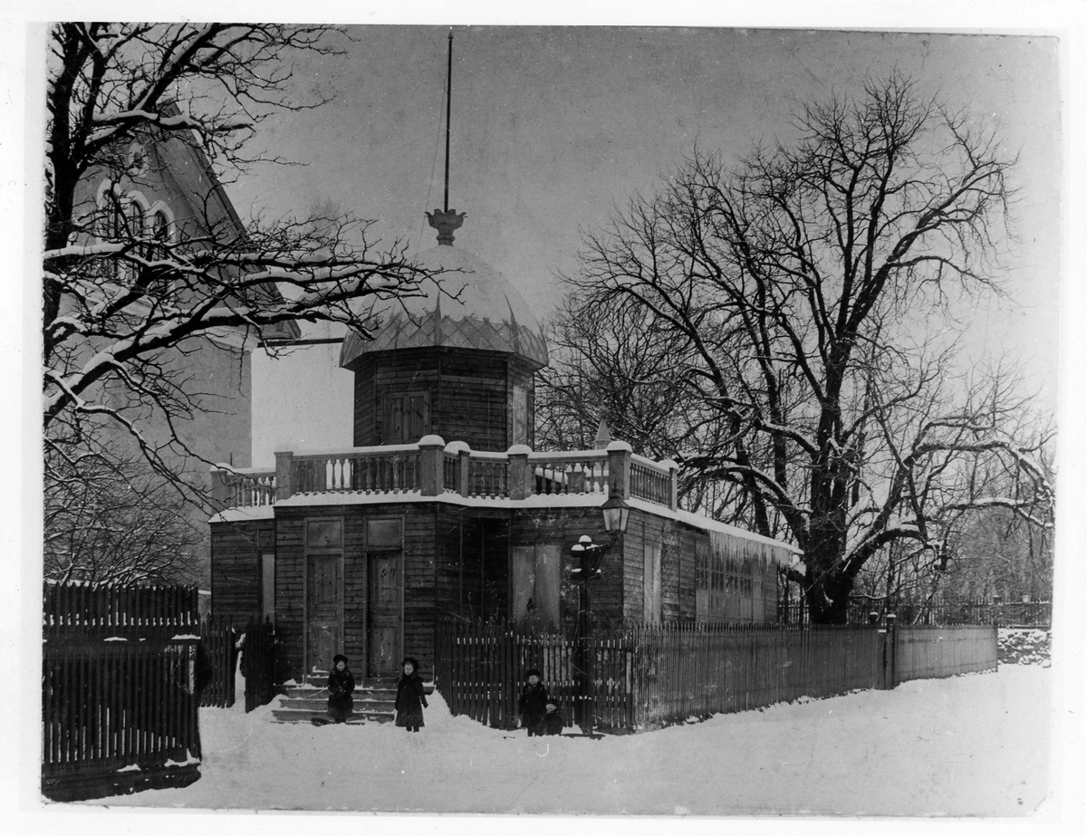 Bild tagen vintertid på Engblads Fotoateljé, en avlång byggnad med en tillbyggd kupol. Ateljén låg vid Alströmska magasinets norra gavel mot Lilla torget. Utanför ingången ses 4 barn. Mark och träd är snötäckta.