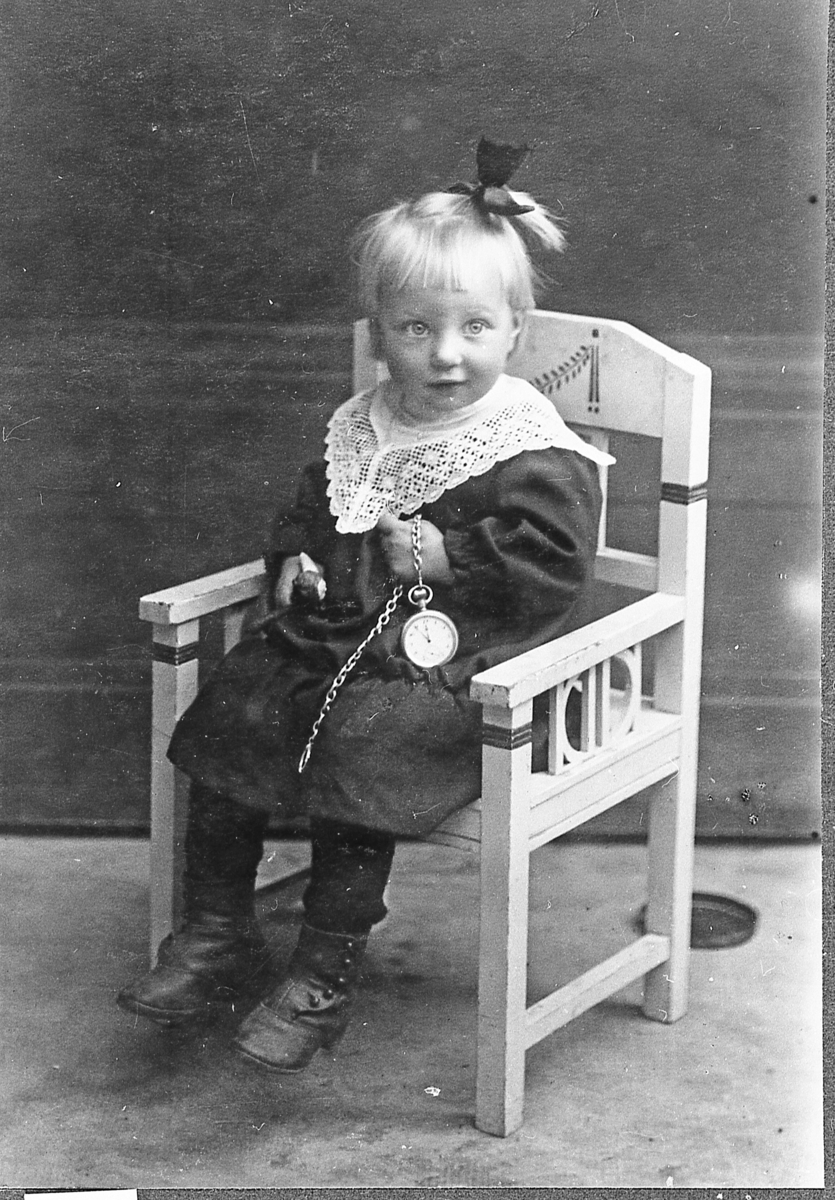 Liten jente med dukke og lommeur, sittende på en stol. Turid Homelien, ca. 1918.