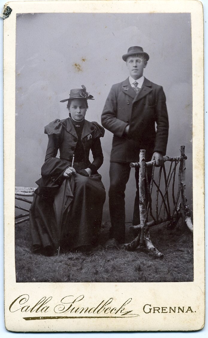 Kabinettsfotografi: gruppbild med en sittande ung kvinna intill en stående ung man. De bär ytterplagg och hatt. Hon håller ett paraply i handen och han håller höger hand innan kavajen, likt Napoleon.