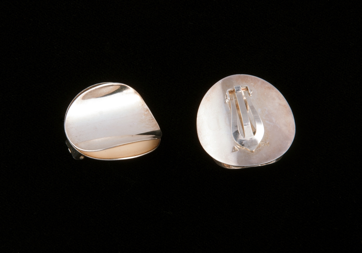 Består av to like store sirkulære sølvringer som er satt sammen langs en midtakse. På hver siden av aksen er ringene bøyd oppover i ulik grad, symmetrisk på hver side.