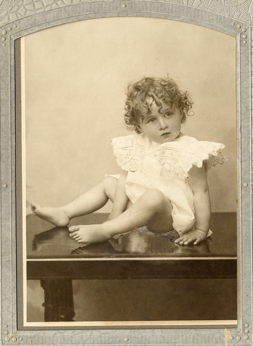 En liten flicka i ljus klänning med brodyr. Hon sitter uppkrupen på ett bord.
Helfigur. Ateljéfoto.

Fotografens dotter.