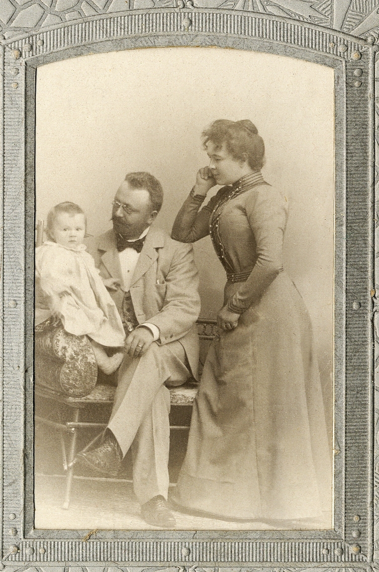 En man i kavajkostym med väst, stärkkrage och fluga, tillsammans med en kvinna i klänning med hög krage. 
De ser på en liten flicka som sitter nära mannen.  
Helfigur. Ateljéfoto.