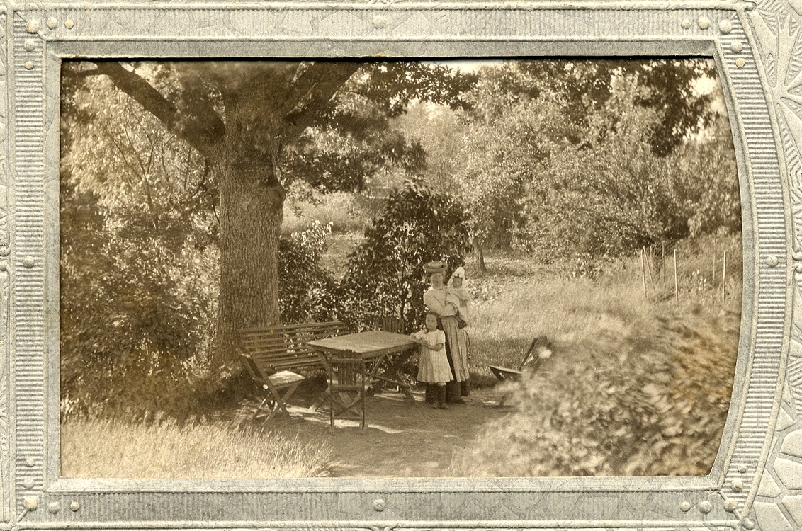 En okänd kvinna i kjol, blus och hatt står vid ett trädgårdsmöblemang i en trädgård, 
tillsammans med två små flickor.