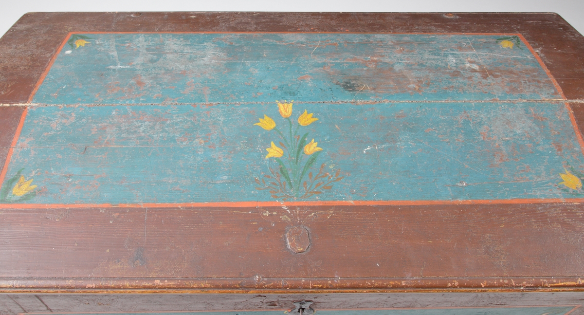 En förvaringskista av trä, brun- och blåmålad med kupat lock. Blomstermålningar runt ägarinitialer:
"APS 1848", på framsidan. Dessa initialer är målade på de ursprungliga ägarinitialerna som är "GLD 1890". På insidan finns ett sidofack med lock.