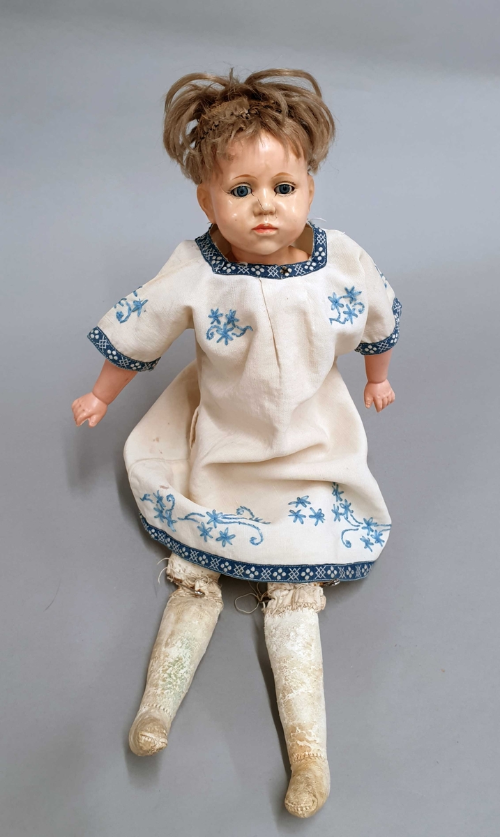 Dukke med øyne som kan lukkes, kort hår og er kledd i en hvit kjole med blå brodering. Hode av keramikk og kropp av skinn. Leggene er festet med sikkerhetsnåler.
