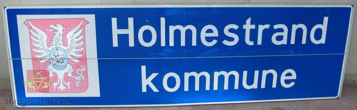 Flerfarget skilt av aluminium der to plater er skrudd sammen med klammer. Skiltet er lysreflekterende, foliert. Med kommunevåpen og har teksten: Holmestrand kommune.