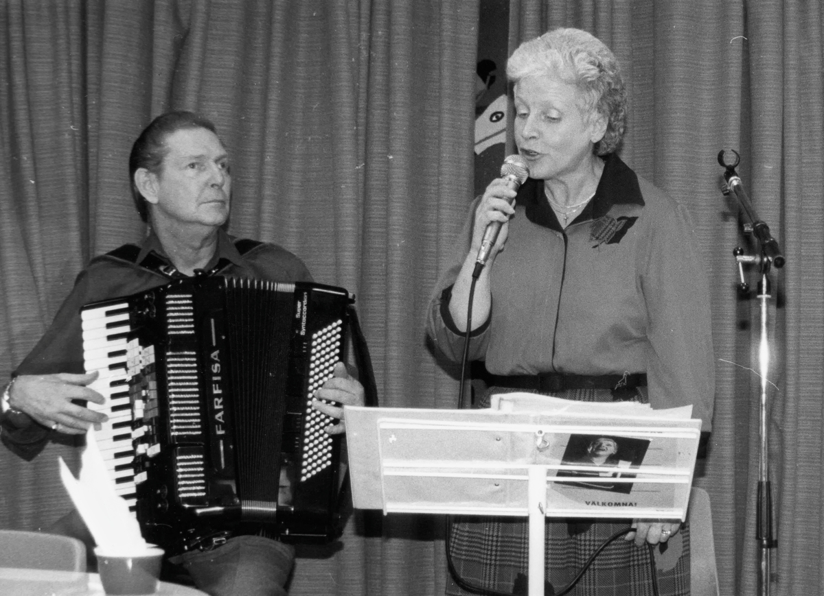 Underhållning vid Kållered hembygdsgilles årsmöte 1986-03-07 i Hallenskolan. En man spelar dragspel och en kvinna sjunger i mikrofon. Namnuppgifter saknas.