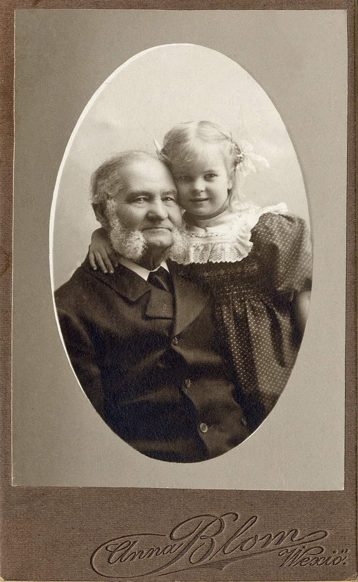 Porträttfoto av en äldre man i mörk kavajkostym m.m.. Troligen tillsammans med sitt barnbarn, en liten flicka i 
mörk klänning och stor, spetskantad krage. 
Bröstbild, halvprofil. Ateljéfoto.