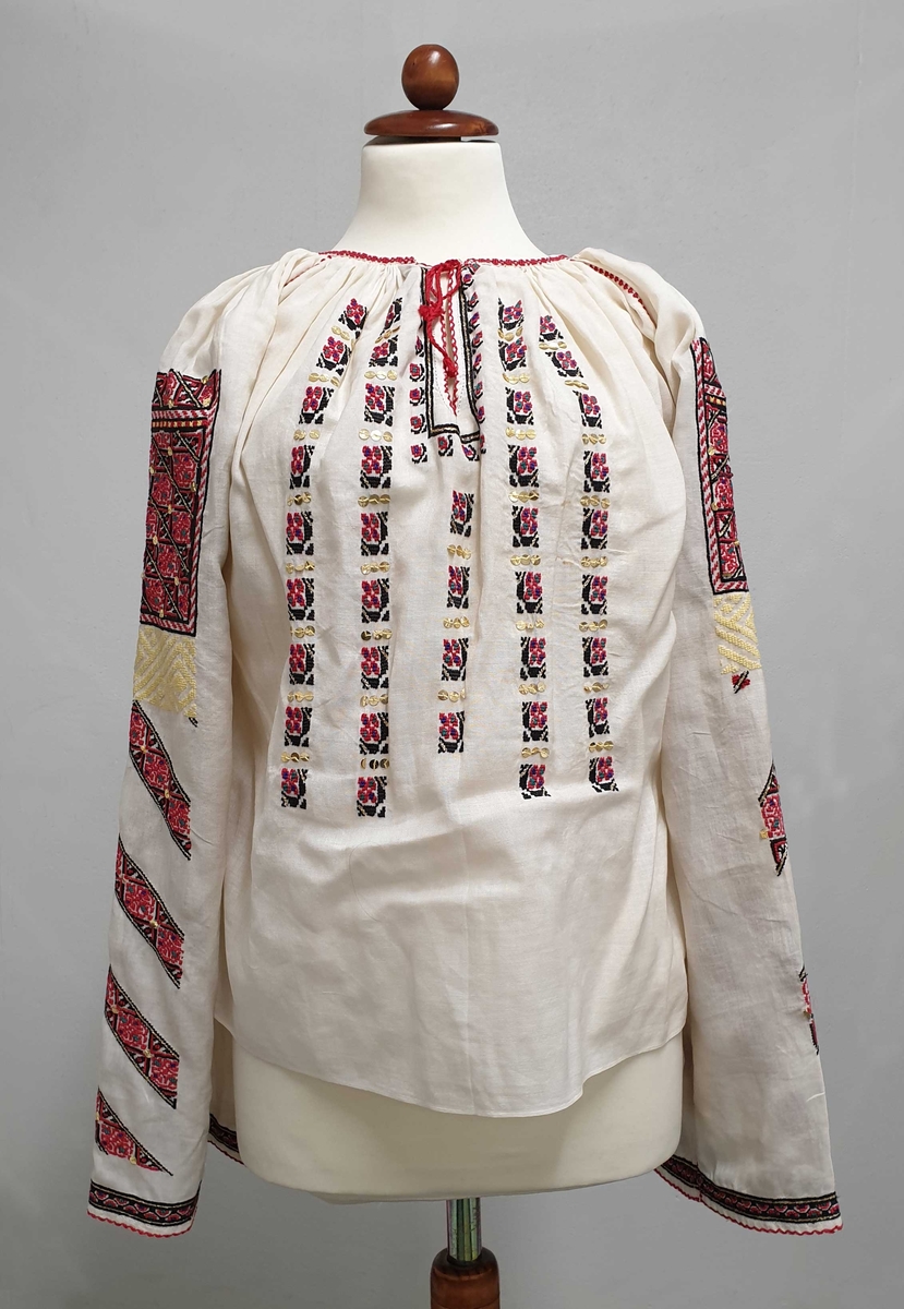 Hvit bluse av lett, nesten transparent bomull, med gullpaljetter og broderier i rødt, svart, gull og gult på bryst og ermer.