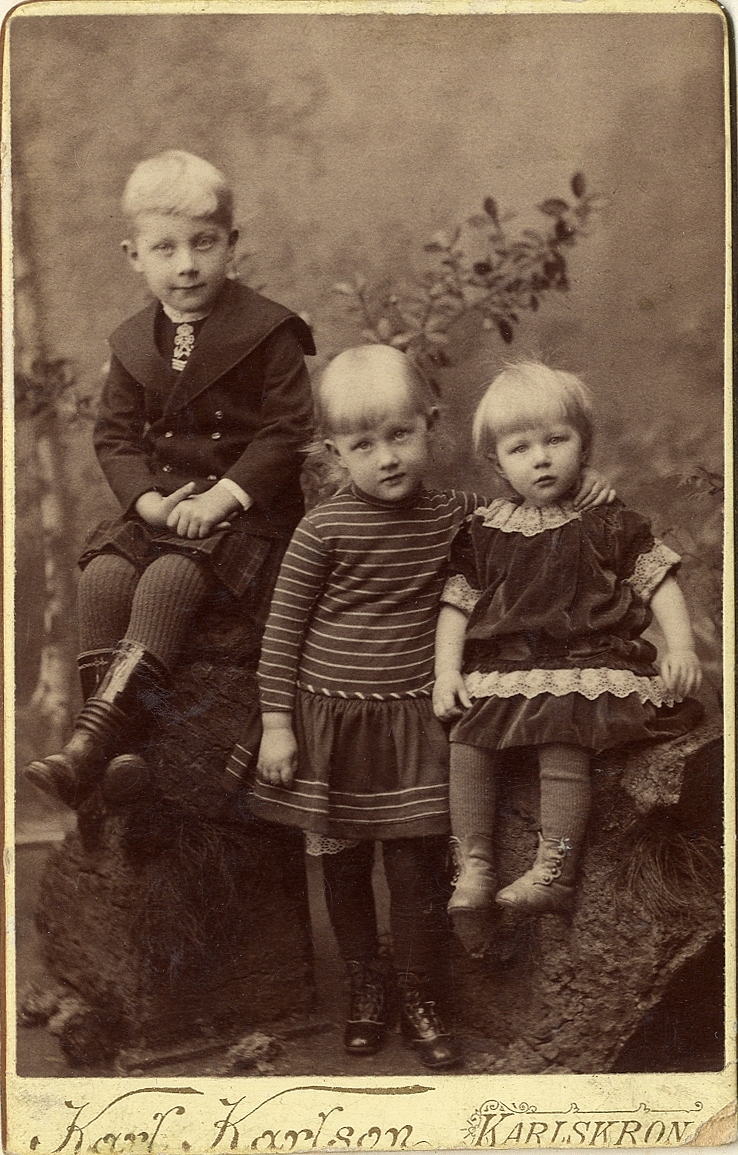 Porträttfoto av tre syskon, en pojke och två flickor. Klädda i diverse barnkläder, bla en sjömansdräkt, en tvärrandig tröja och en sammetsklänning med spetskrage. 
Helfigur. Ateljéfoto.