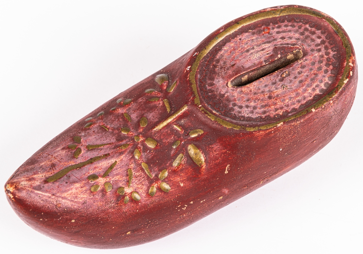 En sparbössa i oljemålat lergods som föreställer en röd träsko/damsko med klack, öppning för mynt och dekorerad med blommönster upptill i guldbronsering. Innuti ligger en liten knapp.