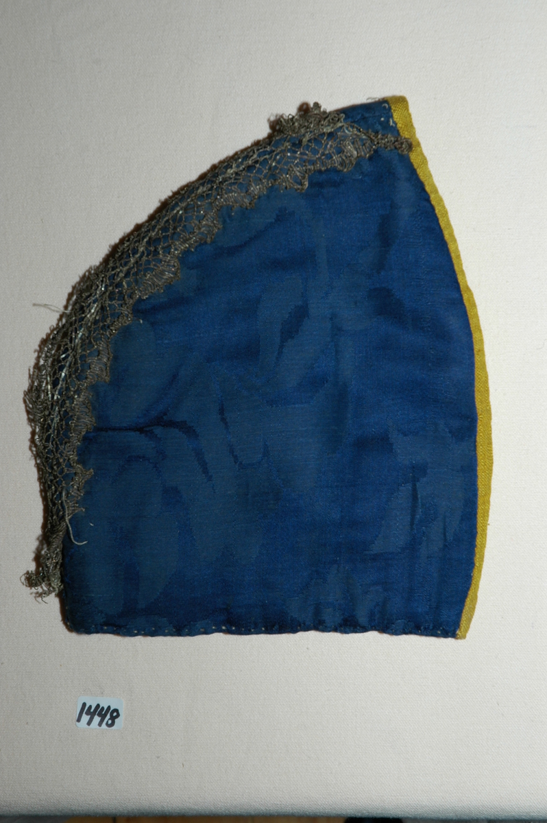 Lue, tobladlue av mørkeblå silkedamask kantet med knallgult silkeband (lerretsvevd). Lua er fôra med linlerret og vattert med stry. Nedrekanten er sydd til med faldesting med grått lingarn. De to delene er sydd sammen med attersting. Sømmonnet er lagt til side og kastet sammen. Kantebandet er vrangsydd på med attersting på retta og faldesting på vranga, 0,5-0,8 cm bredt. Langs sammensyingssømmen er det satt på to rader med en ca. 2 cm brei knipling. Registrering ved Bunad- og folkedraktrådet/Norsk institutt for bunad og folkedrakt v/Jon Fredrik Skauge, 2005, revidert v/Bjørn Sverre Hol Haugen, 2020.