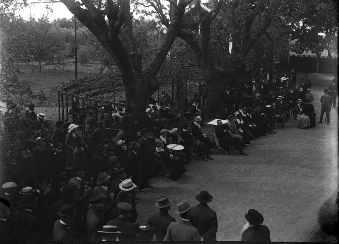 En stor folksamling med stående och sittande i Ribbagårdens trädgård, skuggade av höga träd. De är vända mot någon till höger som inte syns i bild, sannolikt föredragshållare och trädgårdsdirektör G Lind.