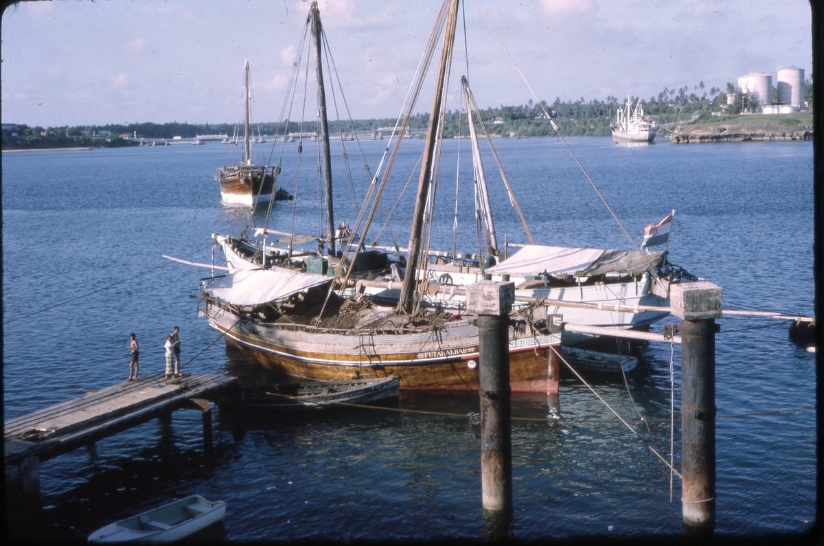 To fortøyde fiskebåter (én med indisk flagg) og joller ved brygge, trolig i Mombasa. 3 mennesker i forgrunnen, og båter og bro i bakgrunnen. 'Sagafjord' Around The World via Africa Cruise 1966.